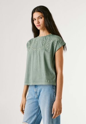 Νεαρή γυναίκα με μακριά σκούρα μαλλιά, που φοράει πράσινο βεραμάν κεντητό μπλουζάκι με κοντά μανίκια και ανοιχτόχρωμο τζιν ψηλόμεσο, όρθια.