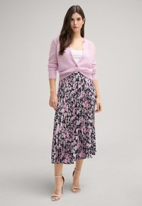 Hellrosane, gestrickte Strickjacke mit Knopfverschluss, über einem floral gemusterten, plissierten schwarzen Rock mit rosa und grünen Akzenten getragen. Kombiniert mit hautfarbenen Sandalen.