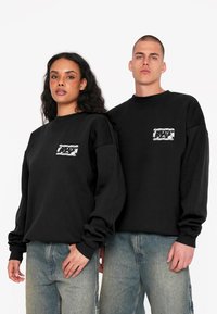 Schwarze Sweatshirts mit Rundhalsausschnitt und abgesetzten Schultern, mit einem grafischen Patch auf der Vorderseite. Kombiniert mit hellblauen Jeans.