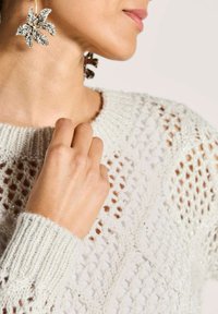 Nahaufnahme einer Frau, die einen cremefarbenen Strickpullover und große funkelnde Blumenohringe trägt, mit ihrer Hand nahe ihrem Hals.