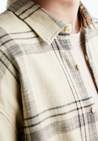 Beige ruudullinen paita nappikauluksella, jossa on mustavalkoiset ruutukuviot, tekstuuri kangasta ja kermaisen väriset napit.
