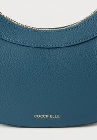 Sininen nahkainen käsilaukku, jossa on kaareva muotoilu, kuvioitu pinta, kullanväriset vetoketjun osat ja kullanvärinen kohokuvioitu logo pohjassa.