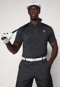 Svart golfpolo med glidelås-krage, kombinert med svarte shorts og et hvitt belte. Modellen holder en golfkølle, iført en hvit caps og hansker.