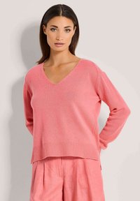 Rosa gestrickter Pullover mit V-Ausschnitt, lässiger Passform, geripptem Saum und langen Ärmeln, getragen mit passenden rosa Leinen-Shorts.