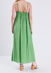 Femme portant une longue robe verte sans manches avec un dos froncé et des sandales à plateforme blanches à talons en bois, debout sur un sol blanc.