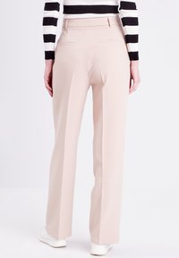 Pantaloni a gamba larga rosa chiaro in tessuto liscio, con tasche posteriori e vita alta, abbinati a sneakers bianche su uno sfondo neutro.