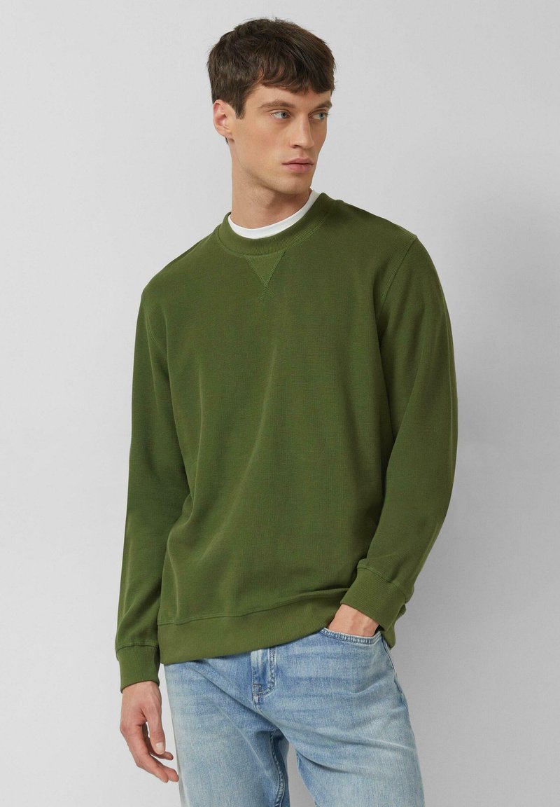 Noor mees, kes kannab oliivirohelist dressipluusi ja helesiniseid teksapükse, seisab ühe käe taskus ja vaatab küljele.