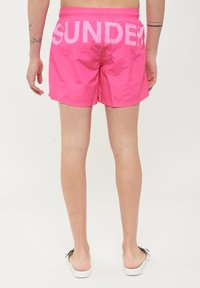 Pantaloni da mare rosa con vita regolabile, caratterizzati da grandi lettere bianche "SUNDECK" sul retro. Tessuto morbido e leggero.