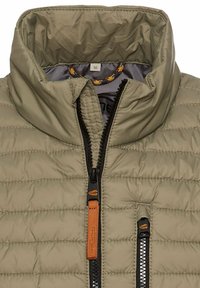 Veste matelassée marron clair avec col montant, fermeture éclair noire et tirette en cuir marron, et poche poitrine zippée avec bordure noire.