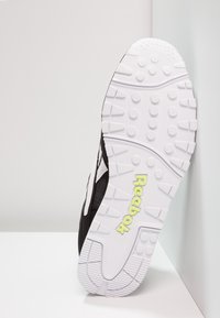 Baskets noires avec semelle extérieure en caoutchouc blanc, dotées d'une semelle texturée et du logo "Reebok" vert. La tige est en tissu avec des accents blancs contrastants.