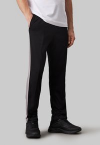 Pantaloni sportivi neri con strisce grigie laterali, realizzati in un tessuto liscio. Presentano due tasche laterali e una vestibilità affusolata. Indossati con sneaker nere.