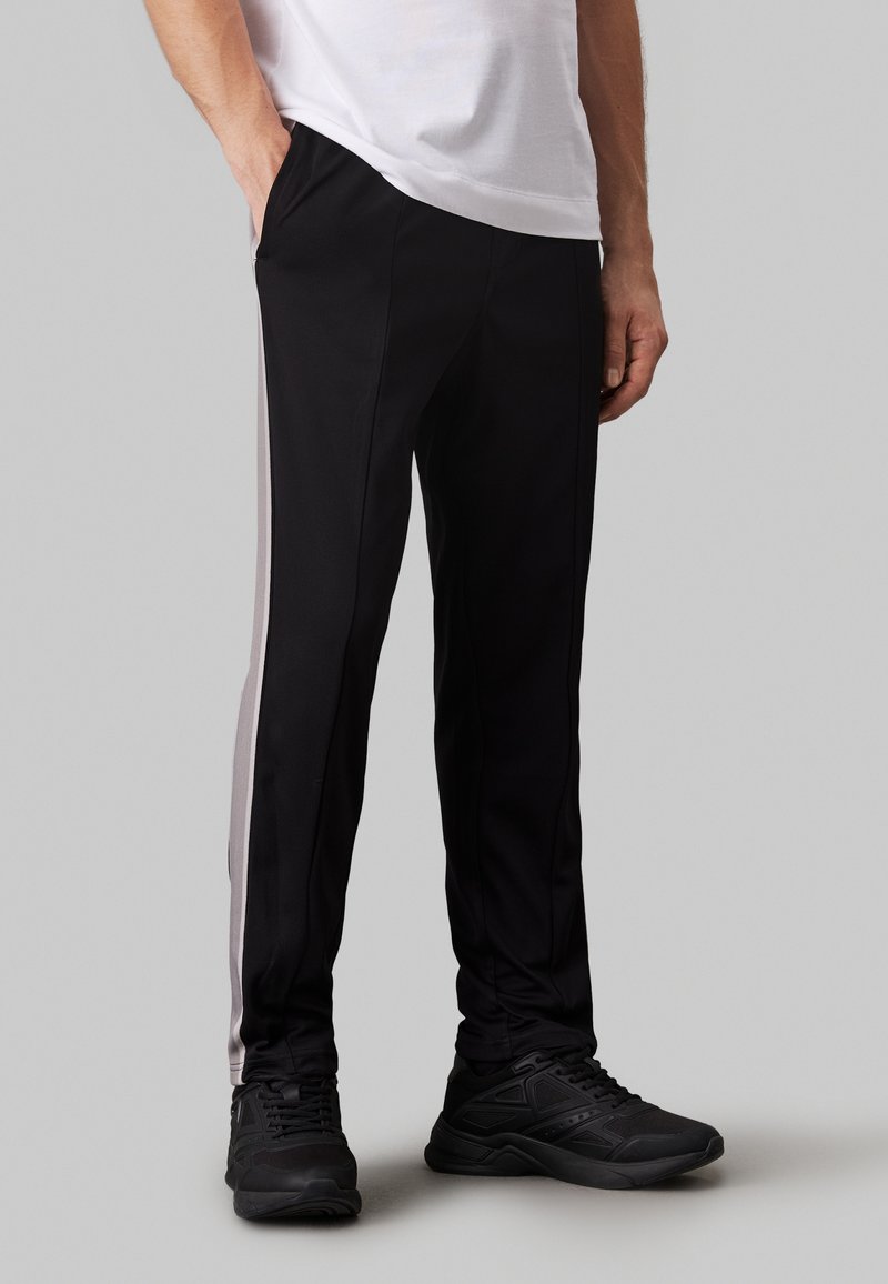 Pantaloni sportivi neri con strisce grigie laterali, realizzati in un tessuto liscio. Presentano due tasche laterali e una vestibilità affusolata. Indossati con sneaker nere.