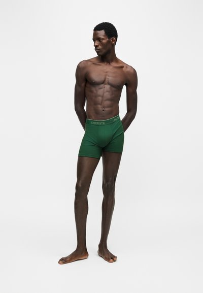 Homme musclé debout pieds nus portant un boxer vert Lacoste, regardant vers sa gauche sur un fond clair uni.