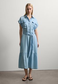 Helles blaues, knielanges Kleid mit kurzen Ärmeln, Knopfverschluss vorne, zwei Brusttaschen und einem gebundenen Taillengürtel; hat einen gestuften Rock.