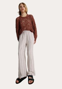 Pull en tricot couleur rouille avec un design texturé, associé à un pantalon large rayé blanc et des sandales noires avec des boucles en argent.