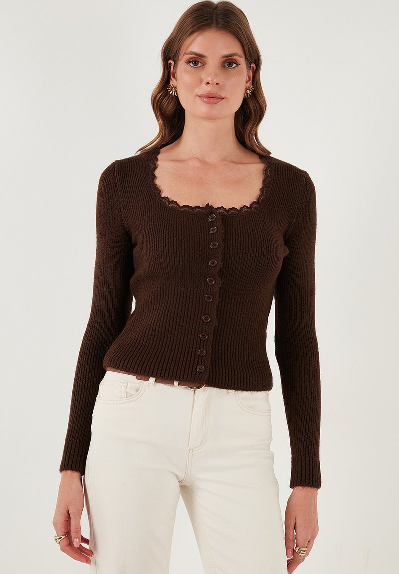 Bruine geribbelde cardigan met een vierkante halslijn, voorzien van een gegolfde rand en acht knopen aan de voorkant. G gedragen met witte jeans.