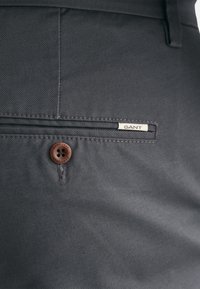Dunkelgraue Hose mit diagonaler Textur, ausgestattet mit einem braunen Knopf und einem GANT-Logoschild auf einer Vordertasche. Gut definierte Nähte und Stickereien.