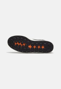 Zwarte rubberen schoenenzool met oranje accenten; voorzien van hexagonale profielpatronen voor grip en een witte midsole voor demping.