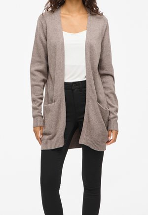 Femme portant un cardigan ouvert beige clair avec poches, une chemise blanche et un pantalon noir, debout devant un fond blanc uni.