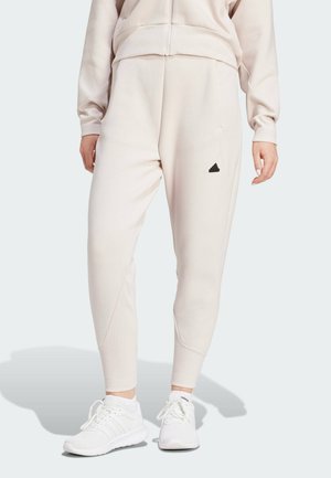 Kvinde iført beige Adidas joggingbukser og matchende zip-up hættetrøje, stående, parret med hvide Adidas sneakers på en ensfarvet baggrund.