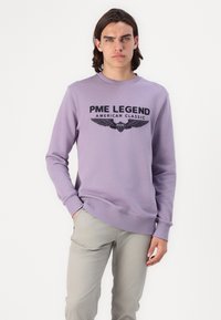 Sudadera morada con cuello redondo, puños y cintura acanalados, que presenta texto negro impreso y un gráfico de alas en la parte delantera. Combinada con pantalones gris claro.