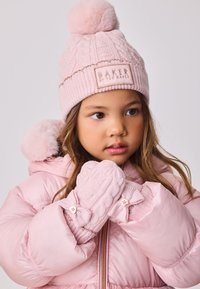 Lyserød strikket hue med pompon og logo-patch; matchende lyserøde vanter med kabelstrik og sløjfedetaljer.