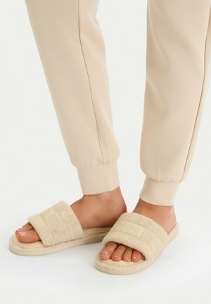 Füße in beigen Fleece-Slides und hellbeigen, umgeschlagenen Jogginghosen vor weißem Hintergrund.