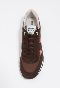 Brązowy zamszowy sneaker z różowymi akcentami, okrągłym noskiem, teksturowanym przodem na sznurowadła i białą gumową podeszwą. Posiada falisty wzór na cholewce.