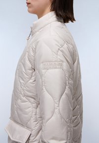 Beige gewatteerde jas met een staande kraag, voorzien van een opvallende logo patch op de mouw en een zijzak voor extra detail.