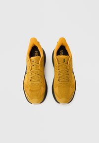 Κίτρινα αθλητικά παπούτσια με υφή πλεκτού πάνω μέρους, ενισχυμένη μύτη και μαύρη εξωτερική σόλα. Διαθέτουν δέσιμο με κορδόνια και επένδυση στον γιακά για άνεση.