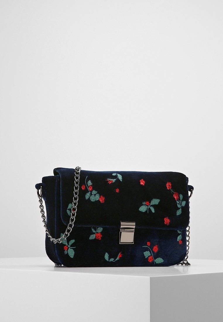 Tmavě modrá sametová kabelka s květinovou výšivkou v červené a zelené barvě, stříbrnými kovovými doplňky a kovovým řetízkem, vybavená magnetickým zapínáním.