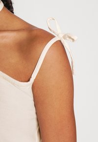 Camisola de cor creme com alças finas amarradas em laços. Tecido suave e leve com um brilho subtil. Apresenta um ajuste descontraído.