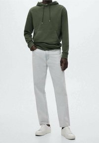 Felpa verde in cotone con coulisse, tasca frontale, abbinata a jeans in denim chiaro. Il modello indossa sneakers bianche. Sfondo semplice.