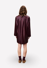 Frau, die mit dem Rücken steht, trägt ein lockeres, langärmliges, dunkelburgunderfarbenes Lederkleid mit Knöchelsocken und flauschigen braunen Hausschuhen.