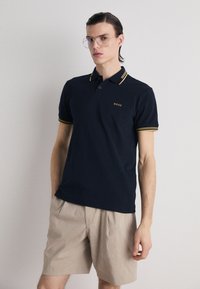 Polo shirt marine avec des accents jaunes, à manches courtes, patte de boutonnage à trois boutons et logo brodé ; associé à un short beige clair.