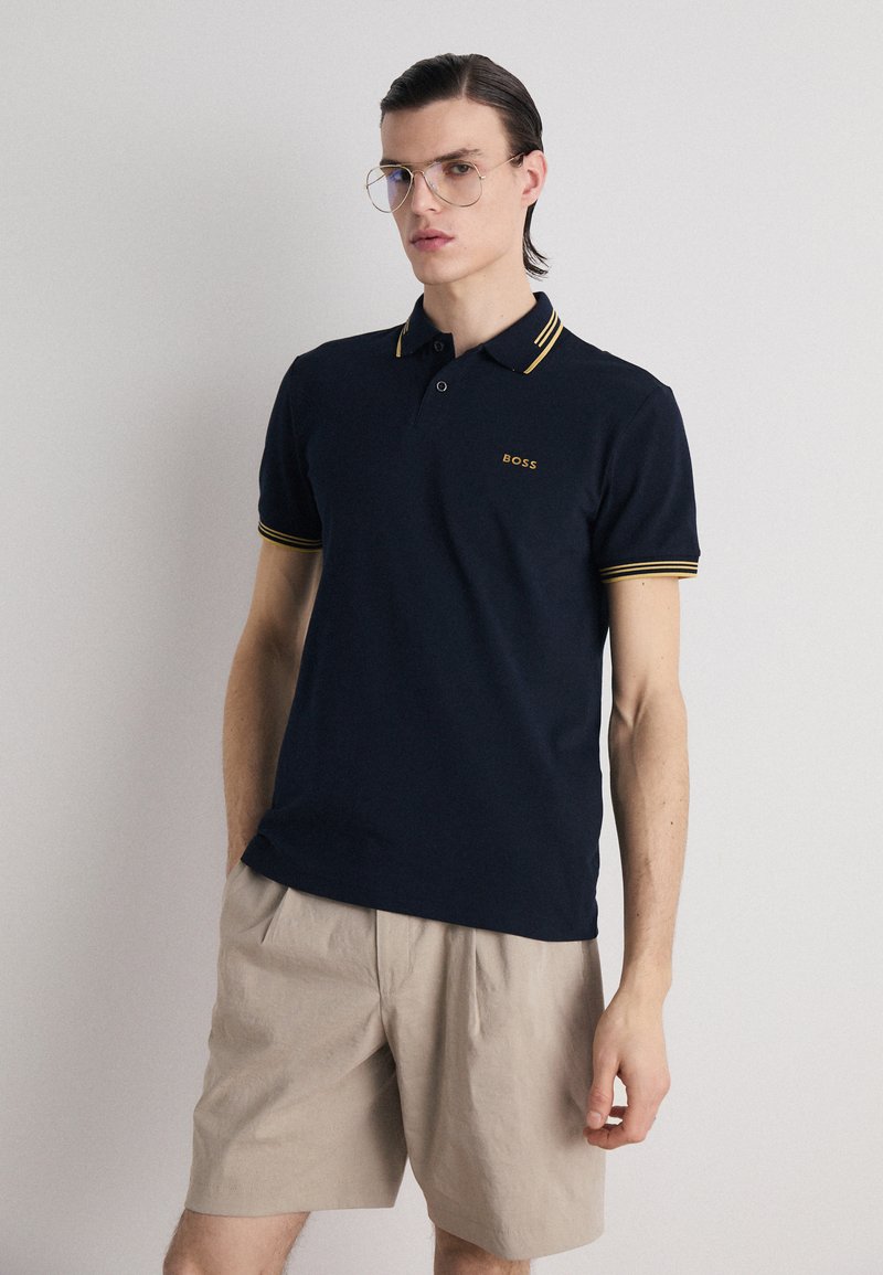 Polo shirt marine avec des accents jaunes, à manches courtes, patte de boutonnage à trois boutons et logo brodé ; associé à un short beige clair.