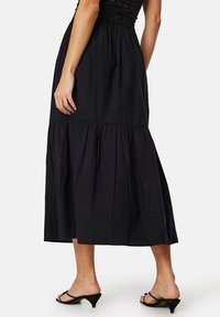 Gonna maxi nera con vita arricciata, design a strati e tessuto leggero. Un paio di sandali neri con tacco visibili nella parte inferiore.