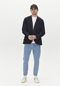 Blazer navy con bolsillo de parche, camiseta blanca, jeans azul claro y zapatillas blancas. Los accesorios incluyen una gorra negra con texto.