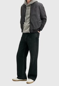 Hombre que lleva una sudadera gris debajo de una chaqueta bomber negra, pantalones vaqueros negros holgados y zapatillas blancas con suelas de goma, con las manos en los bolsillos.