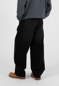 Pantaloni in denim nero dalla vestibilità ampia, con due tasche posteriori e cuciture a contrasto. Indossati con scarpe marroni senza lacci su uno sfondo semplice.