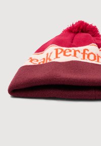 Tumši sarkans adīts cepures modelis ar noslēgtu maliņu, kuram ir balta un oranža logo lente, kā arī pom-poms augšpusē. Mīksta tekstūra, silts materiāls.