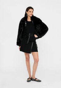 Črni bomber jakna iz umetnega krzna z velikimi rokavi, združena s črnim topom in asimetrično krilo, dopolnjena s črnimi ravnimi čevlji.