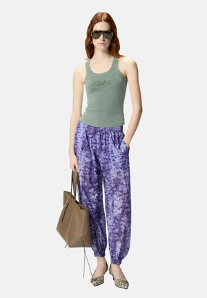 Donna che indossa una canotta verde, pantaloni viola con fantasia, tiene una borsa marrone, indossa occhiali da sole e ballerine a punta in pelle di serpente, in piedi.