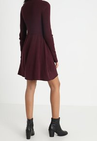 Femme portant une robe rouge bordeaux à manches longues et à motif côtelé, avec des bottines noires, se tenant contre un fond blanc uni, le dos tourné vers l'avant.