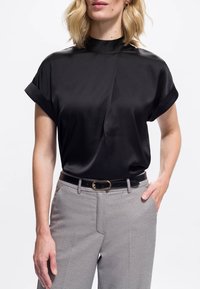 Femme portant un blouse en satin noir à manches courtes rentrée dans un pantalon gris à motifs taille haute avec une ceinture noire, se tenant avec une main dans la poche.