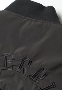 Veste bomber noire avec un col côtelé et un texte brodé en fil noir structuré au dos. Surface en tissu lisse et brillant.