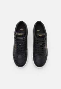 Černé kožené tenisky s hladkým texturem, se perforovanými prvky, plochými černými tkaničkami a zlatým logem "BOSS" na jazyku. Bílé podešev.