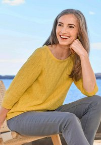 Gelber Strickpullover mit einem strukturierten Muster, rundem Ausschnitt und Dreiviertelärmeln, kombiniert mit grauen Hosen. Lässige Außenszenerie.