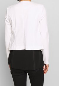 Vit kort jacka med lång ärm, rena linjer och en slät textur, i kombination med en svart lagerblus under.