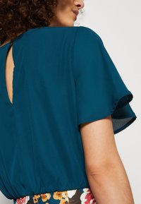 Tee-farbene Bluse mit kurzen, ausgestellten Ärmeln, elastischem Bund und einem Schlüsselloch-Detail im Rücken. Der Stoff wirkt glatt und leicht und wird von einem blumigen Rock akzentuiert.